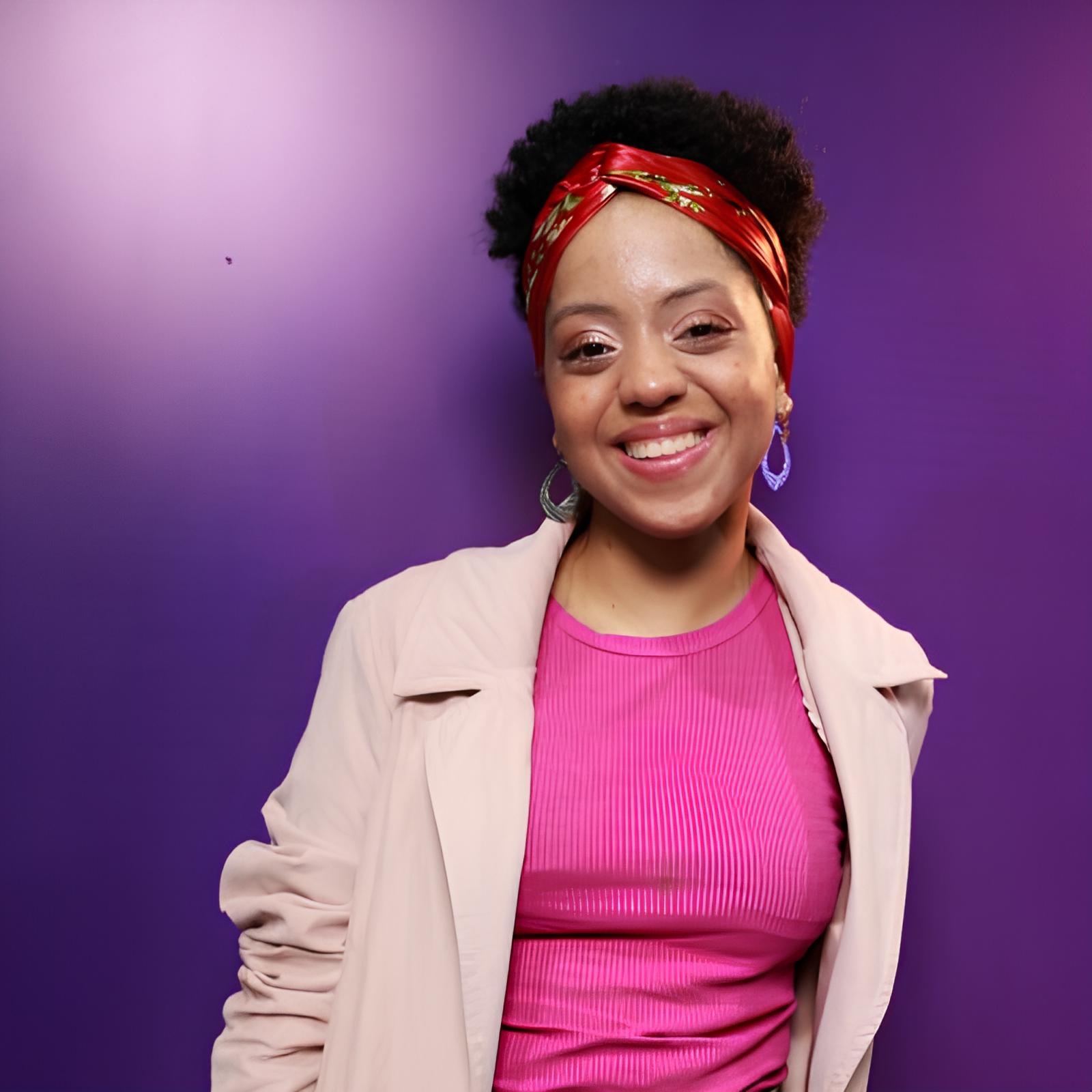 Foto de Larissa Souza, mulher negra com cabelo crespo peso, está vestindo camiseta roxo e um sobretudo rose
